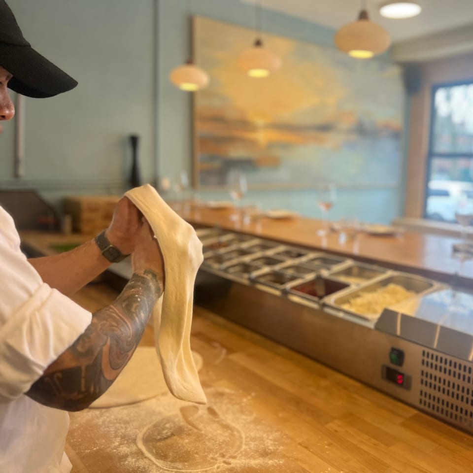 A chef with tattooed arms stretches pizza dough at One World Pizza's counter.