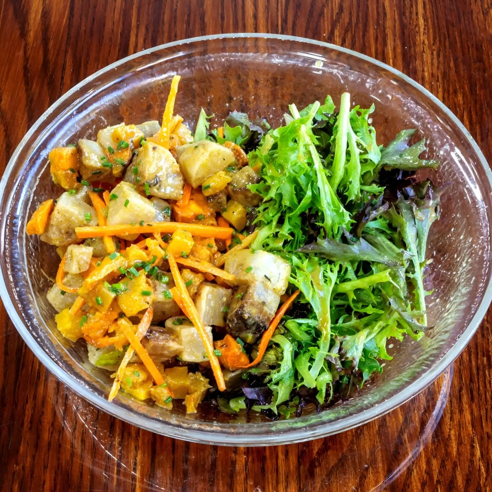 A glass bowl of roasted potato and carrot salad with mixed greens on a wooden table.