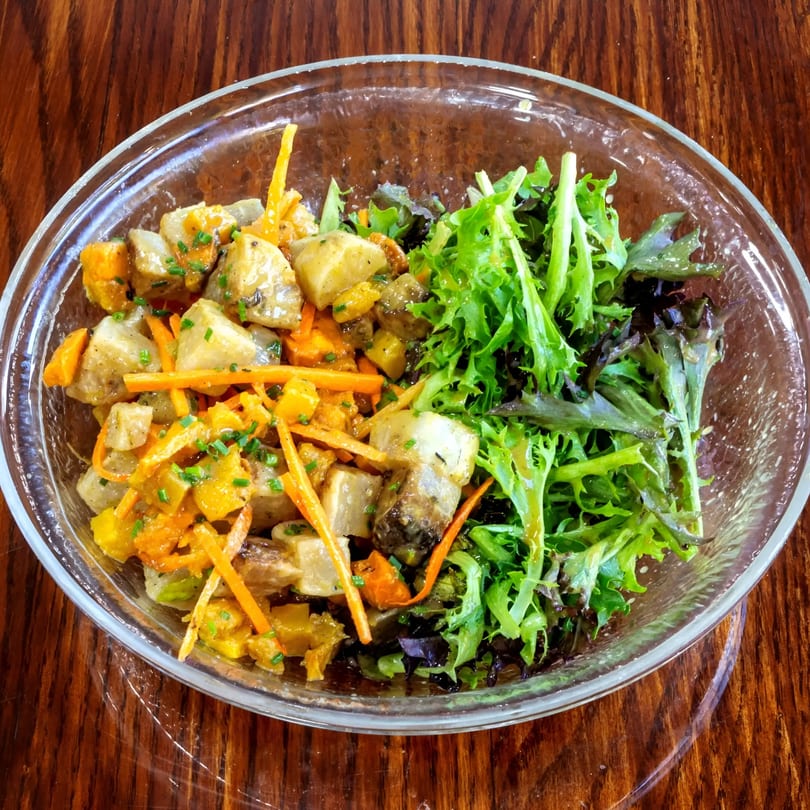 A glass bowl of roasted potato and carrot salad with mixed greens on a wooden table.