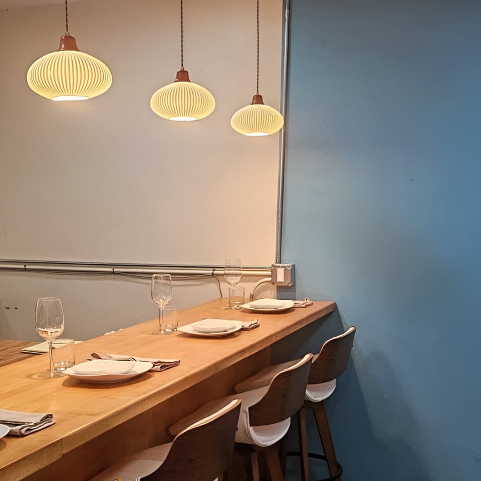 A long wooden bar with place settings and three modern bar stools under hanging pendant lights.