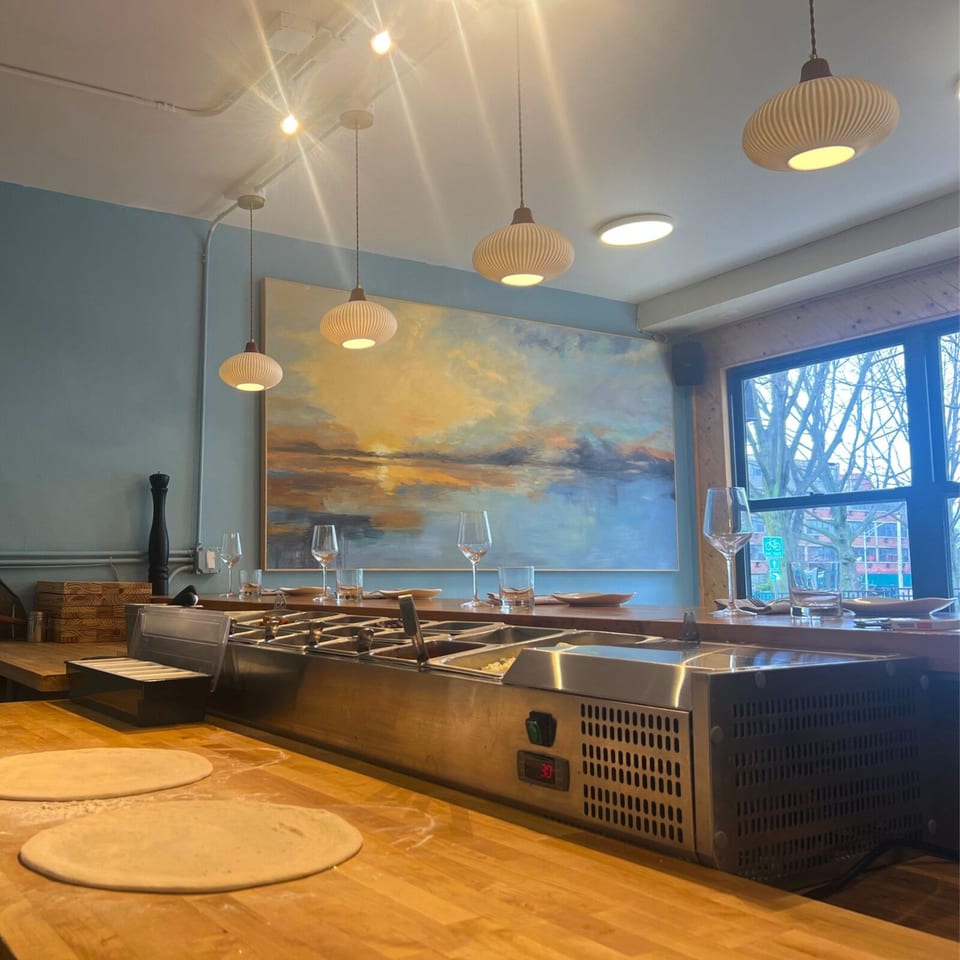 Interior of One World Pizza with pizza dough on a wooden counter, ingredient bar, and abstract art.