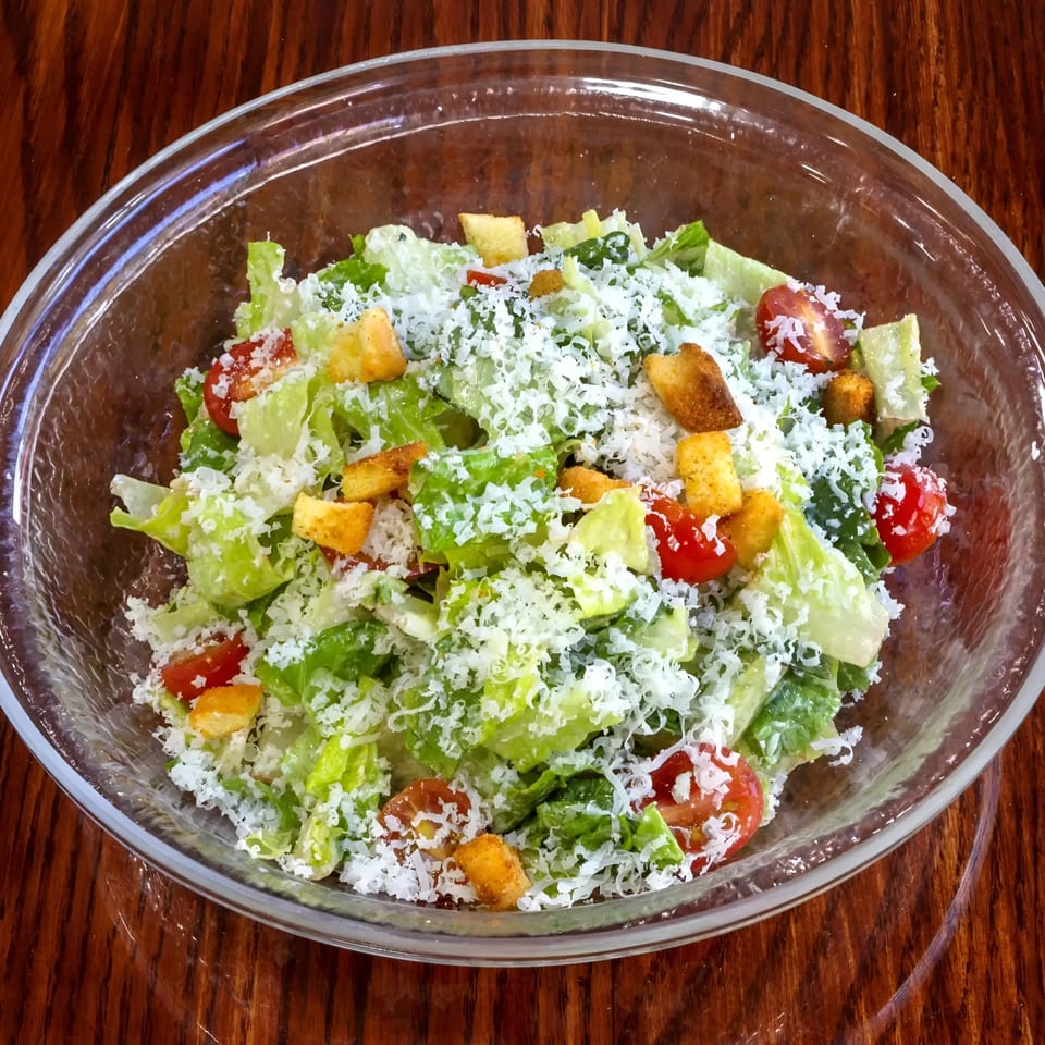 A Caesar salad in a clear bowl, topped with shredded cheese, croutons, and cherry tomatoes.